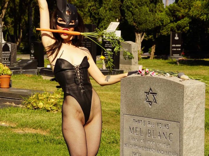 a woman in a bunny mask and lingerie poses next to a gravestone with a carrot in her mouth