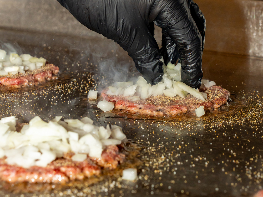 Three burger patties being pressed to a plancha with onions on top