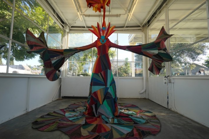A colorful robe sculpture with arms outstretched and a feathered headdress in a space of glass windows.