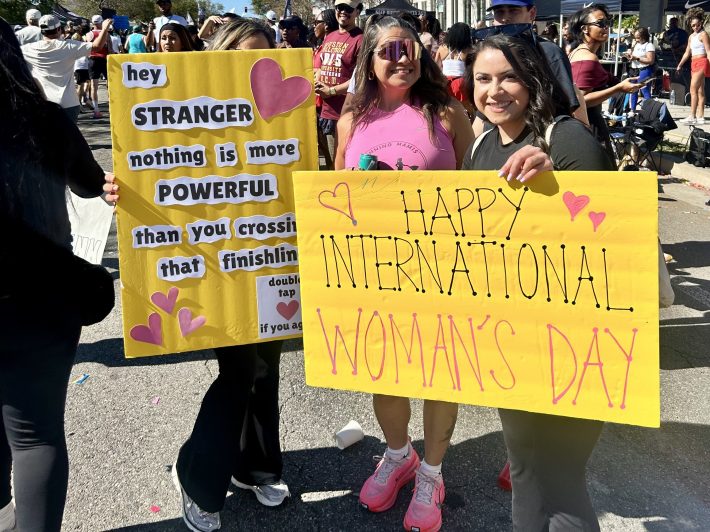 ladies with signs saying "happy international women's day" and "hey stranger, nothing is more powerful than you crossing that finish line"