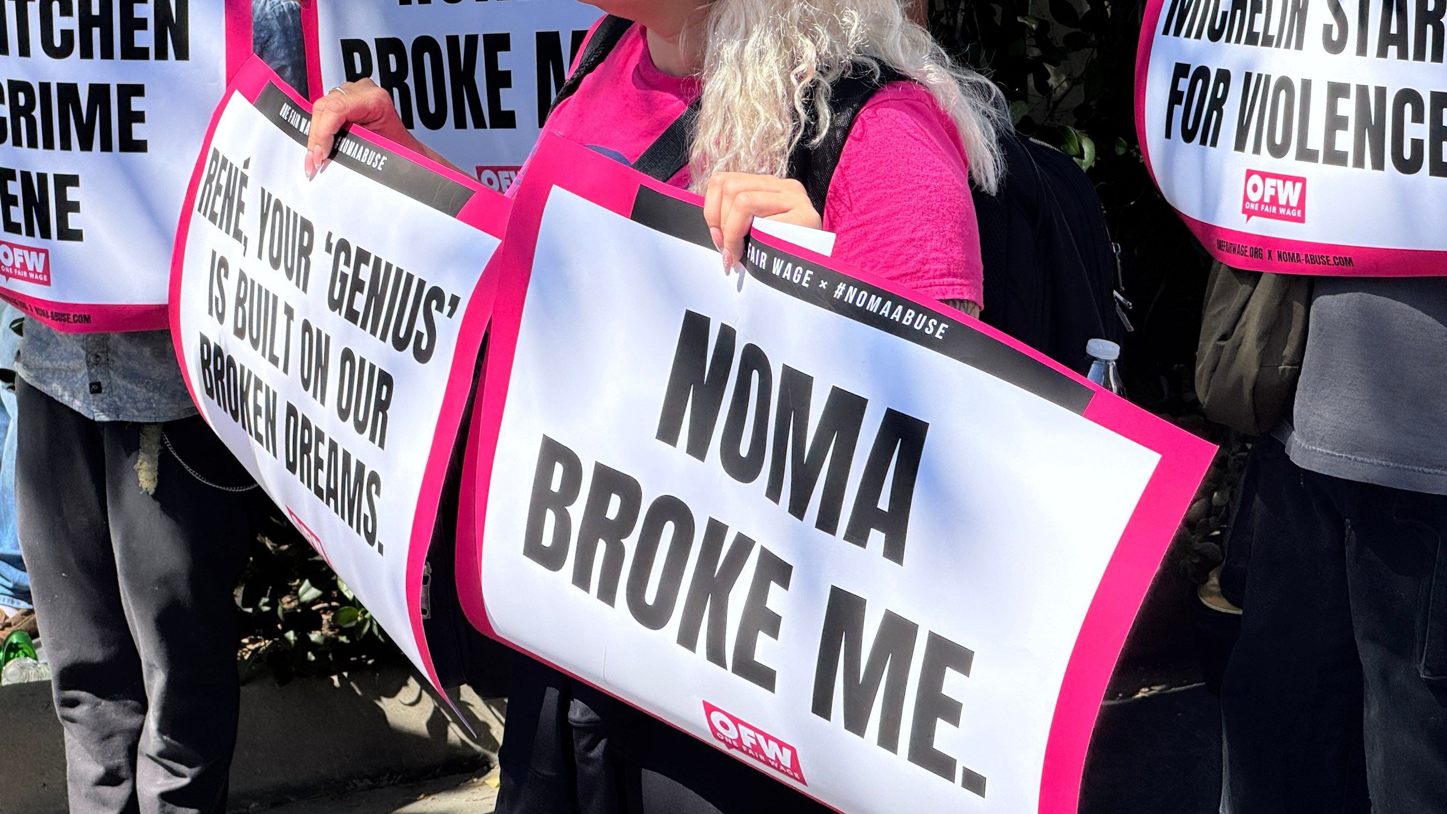 An organizer with One Fair Wage holds a sign that says "NOMA BROKE ME" in front of the Paramour Estate in Silver Lake