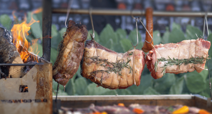 Rosemary-tied meats hanging from hooks over an open grill next to a flaming log