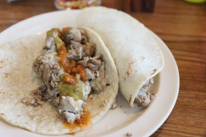 Two tacos, one open, showing cooked cow brains and swabs of red and green salsa, next to one that is rolled up