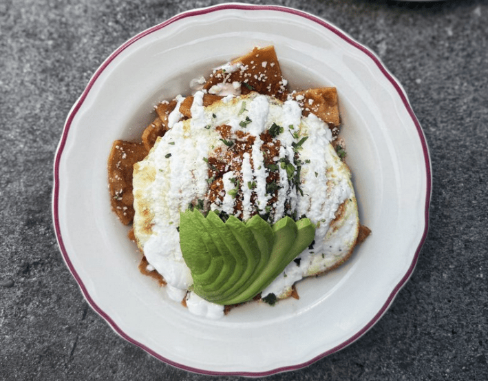 A white plate with chilaquiles, topped with egg and avocado, salsa and crema
