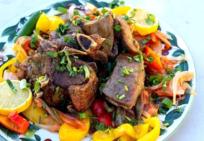 A salad topped with fried beef and yellow bell pepper strips, sprinkled with a green herb and a wedge of lemon on the right side