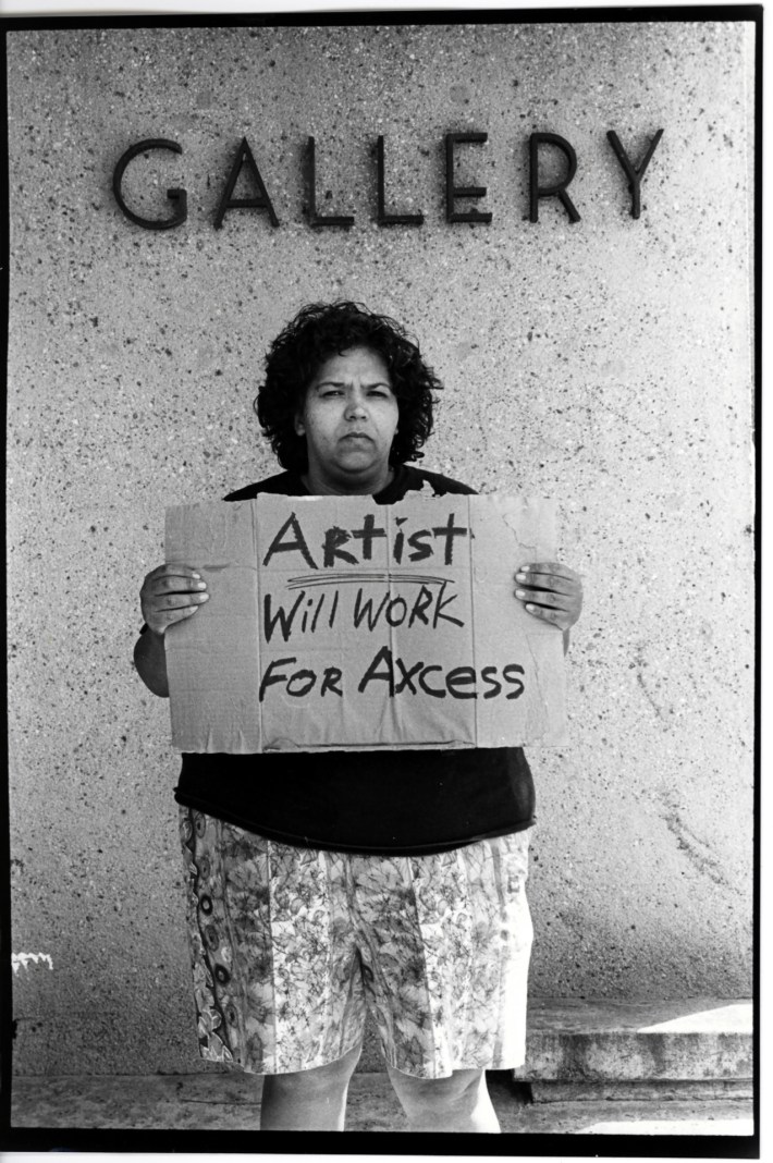 A black-and-white photograph of artist Laura Aguilar holding a cardboard sign that says "ARTIST WILL WORK FOR AXCESS" while standing in front of a "gallery" sign