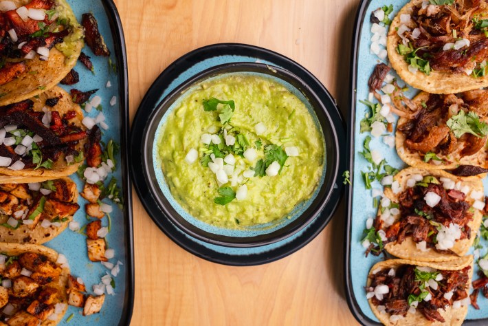 A small bowl of guacamole topped with onions and cilantro between two trays of tacos on corn tortillas