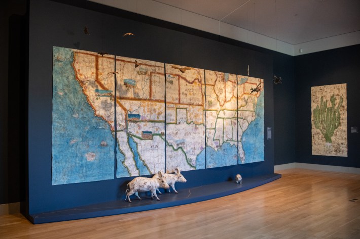"Book 13" installation by Sandy Rodriguez at The Huntington. The installation features a 20-foot-wide painted U.S. map with states outlined in orange, yellow, and green to signify if they were free or slave states. 3D elements include life-size pigs and a miniature hanging helicopters.