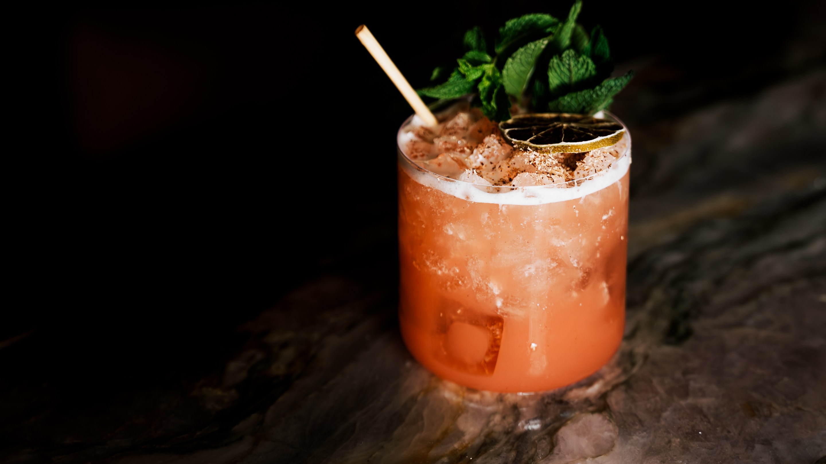 A reddish Junglebird cocktail with a straw, dried citrus rind, and mint