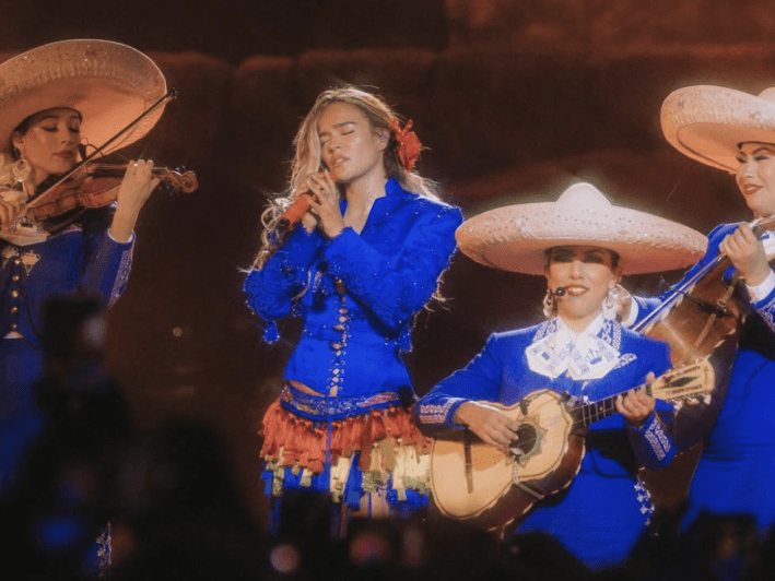 a woman sings onstage with women mariachi performers around her
