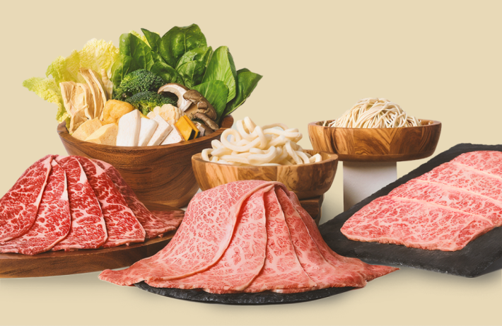 Three kinds of raw Wagyu beef on black trays, backed by bowls of noodles, tofu, vegetables, and lettuce