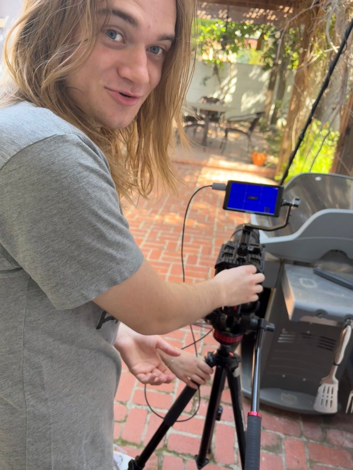 Collins stands behind a video camera mounted on a tripod looking at the camera with a smile on his face.
