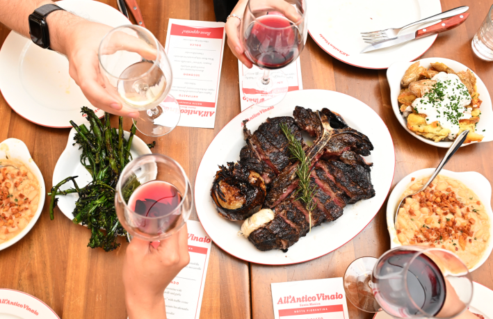 A table set with pasta dishes, and a giant t-bone steak in the center, ringed with four hands holding glasses of wine