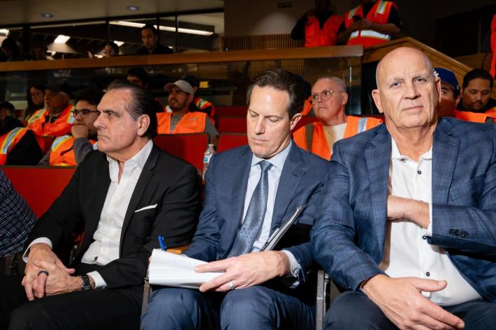 three men in suits and men in orange vests behind them