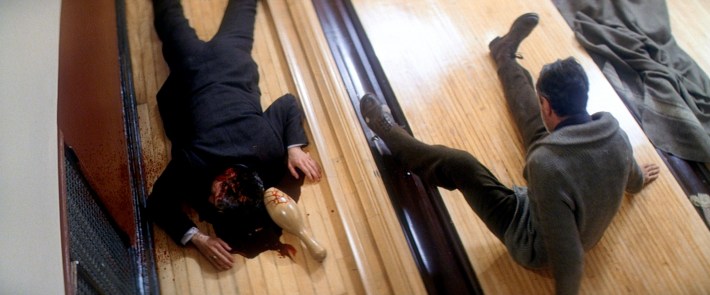 a character lays dead, bludgeoned to death by a bowling pin next to a man in a suit