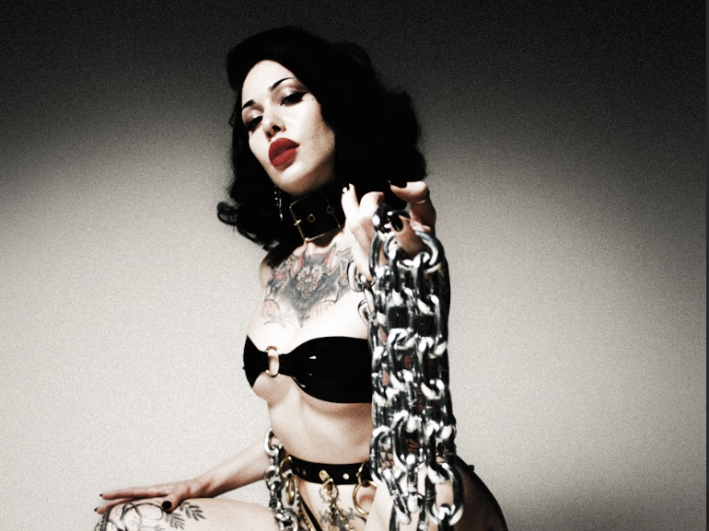 a domme wears lingerie and holds a large chain while squatting