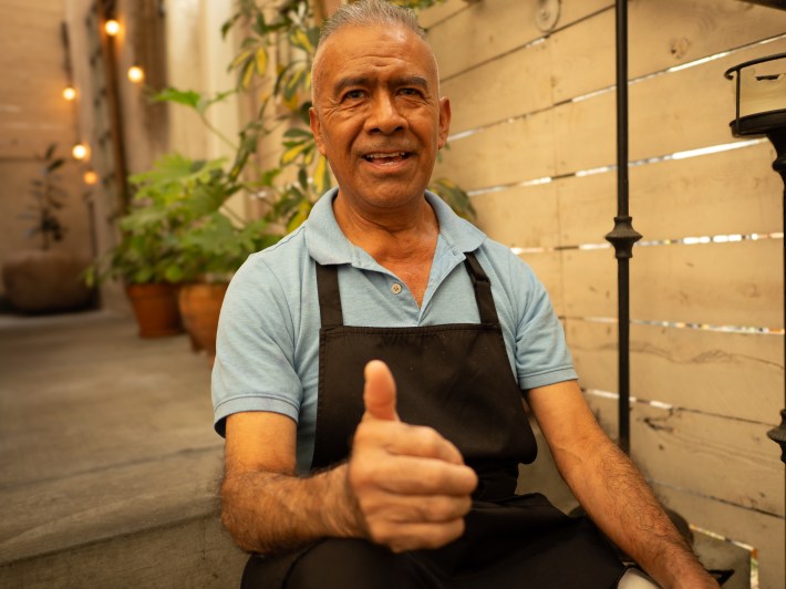 a man in an apron and blue polo gives a thumbs up