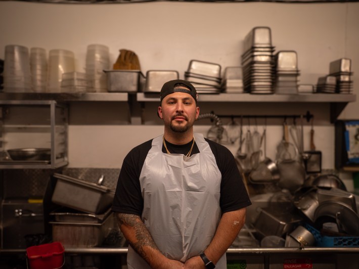 a man in a cap wears an apron in front of a sink with many silver tubs and containers
