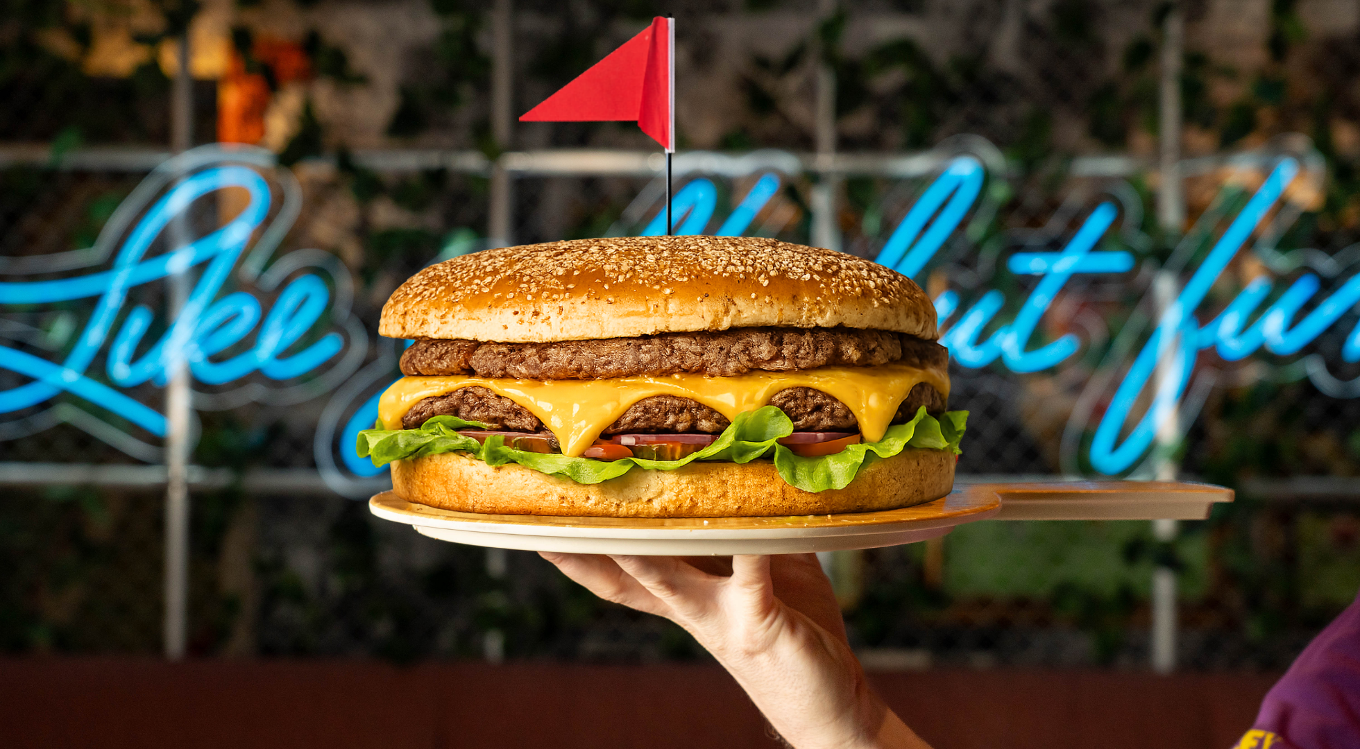 A giant five pound burger with a red flag in the top, being held by a hand as it sits on a tray