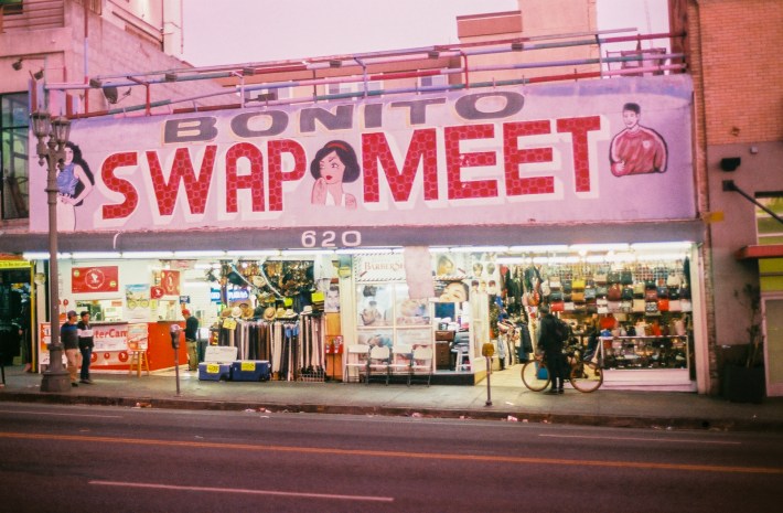 People walk past the Bonito Swap Meet sign. Inside the swat meet, shops are open.