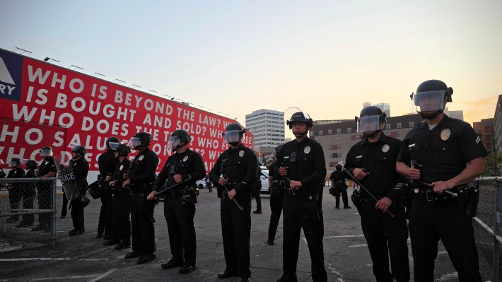 A line of officers wearing riot helmets and holding batons in front of a giant mural.