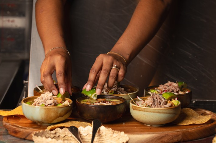 A woman's hands plating two bowls of pozole, with other bowls around her.