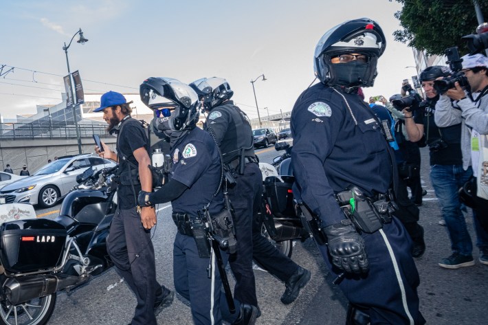 LAPD motorcycle officers grab journalist's arms and escort him behind police line.