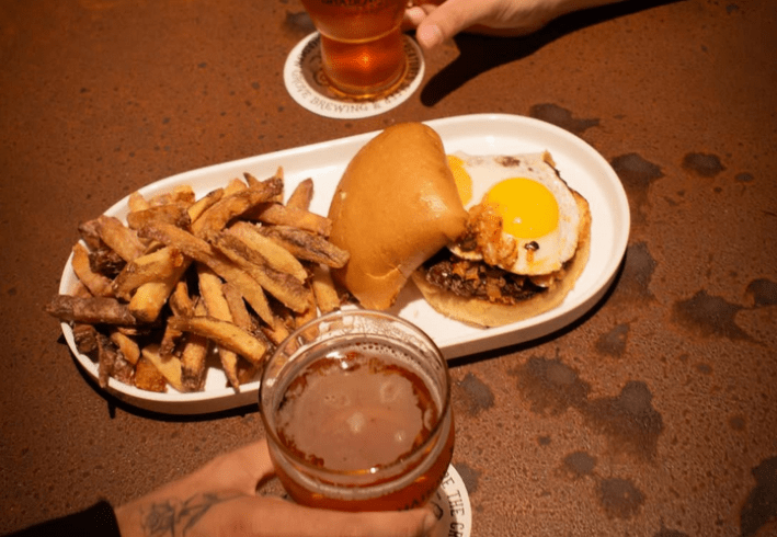 A white tray with a burger and fries. The burger has two sunny side up eggs on top, while hands hold beers on the periphery