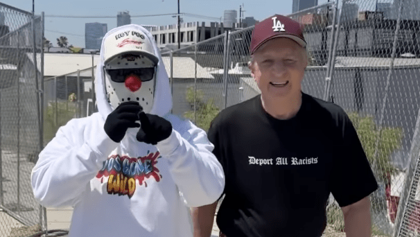a man in a mask and hoodie walks beside a man in a LA cap