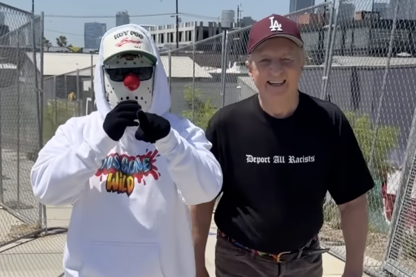 a man in a mask and hoodie walks beside a man in a LA cap