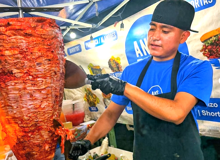 A taquero with a blue shirt and backward baseball cap slicing meat from a giant trompo of al pastor.