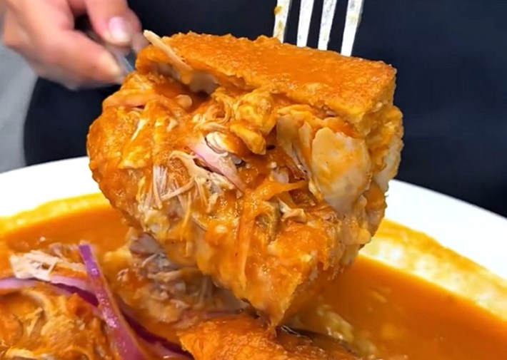 A saucy torta ahogada being lifted by two forks over a white plate of red sauce