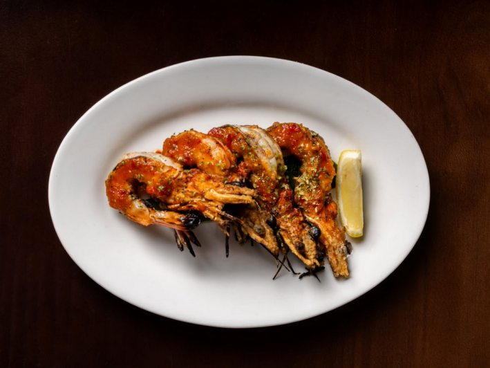 Four grilled blue prawns with Balinese Jimbaran tomato glaze on a white plate with a wedge of lemon
