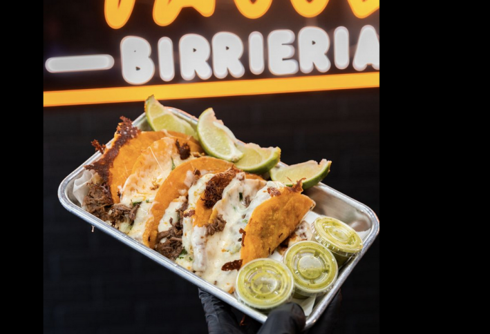 3 quesabirria tacos on a metal tray with three plastic cups of green salsa and lime wedges on the side, all infront of  asign that says "birrieria"