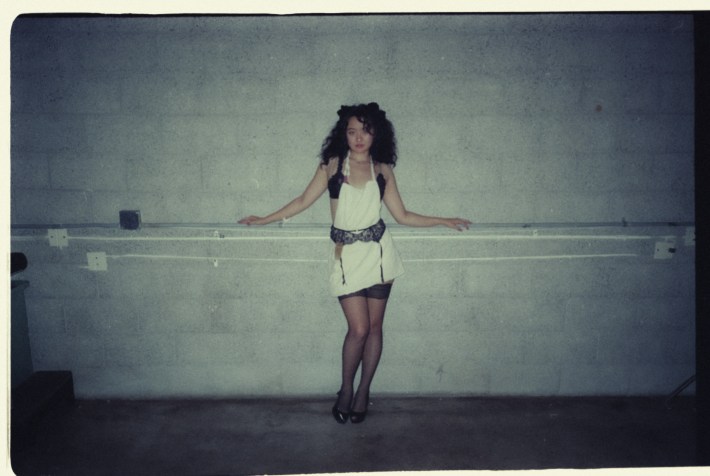 A woman in a French maid outfit stands with her arms spread against a brick wall