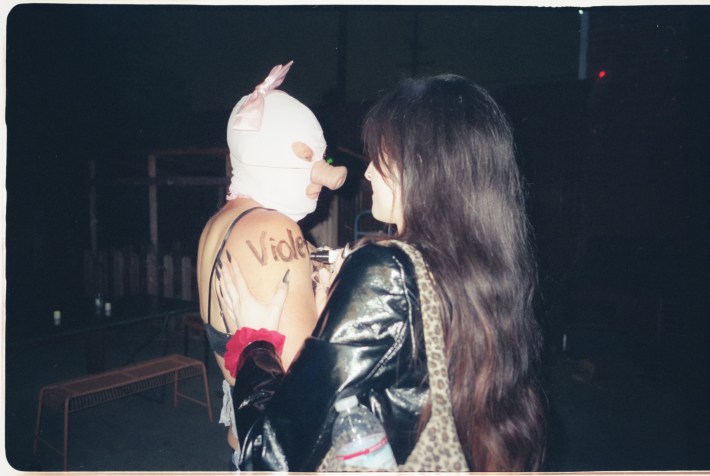 A woman writes her name on a man in a pig mask, as seen from behind her