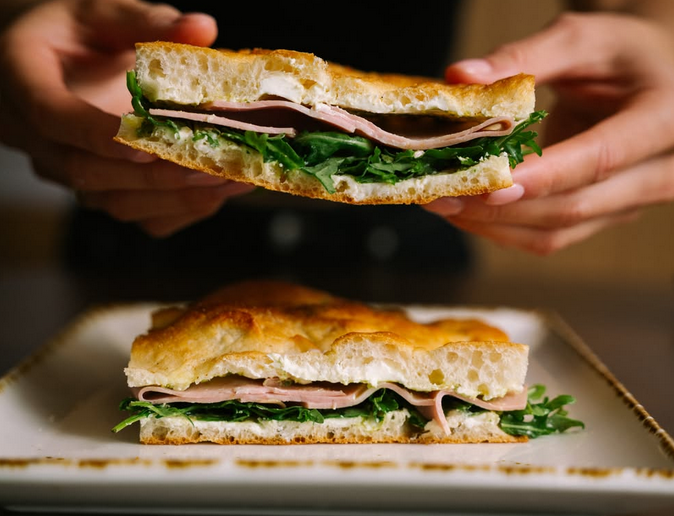 A mortadella sandwich with mortadella, pesto, and mozzarella on fresh focaccia., one of the halves being held up by someone's hands.