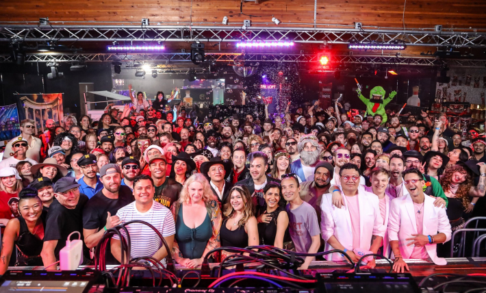 A photo of a lot of people at a party, under overhear lighting tracks. In back is Slimer from Ghostbusters. One attendee wears a fake beard.