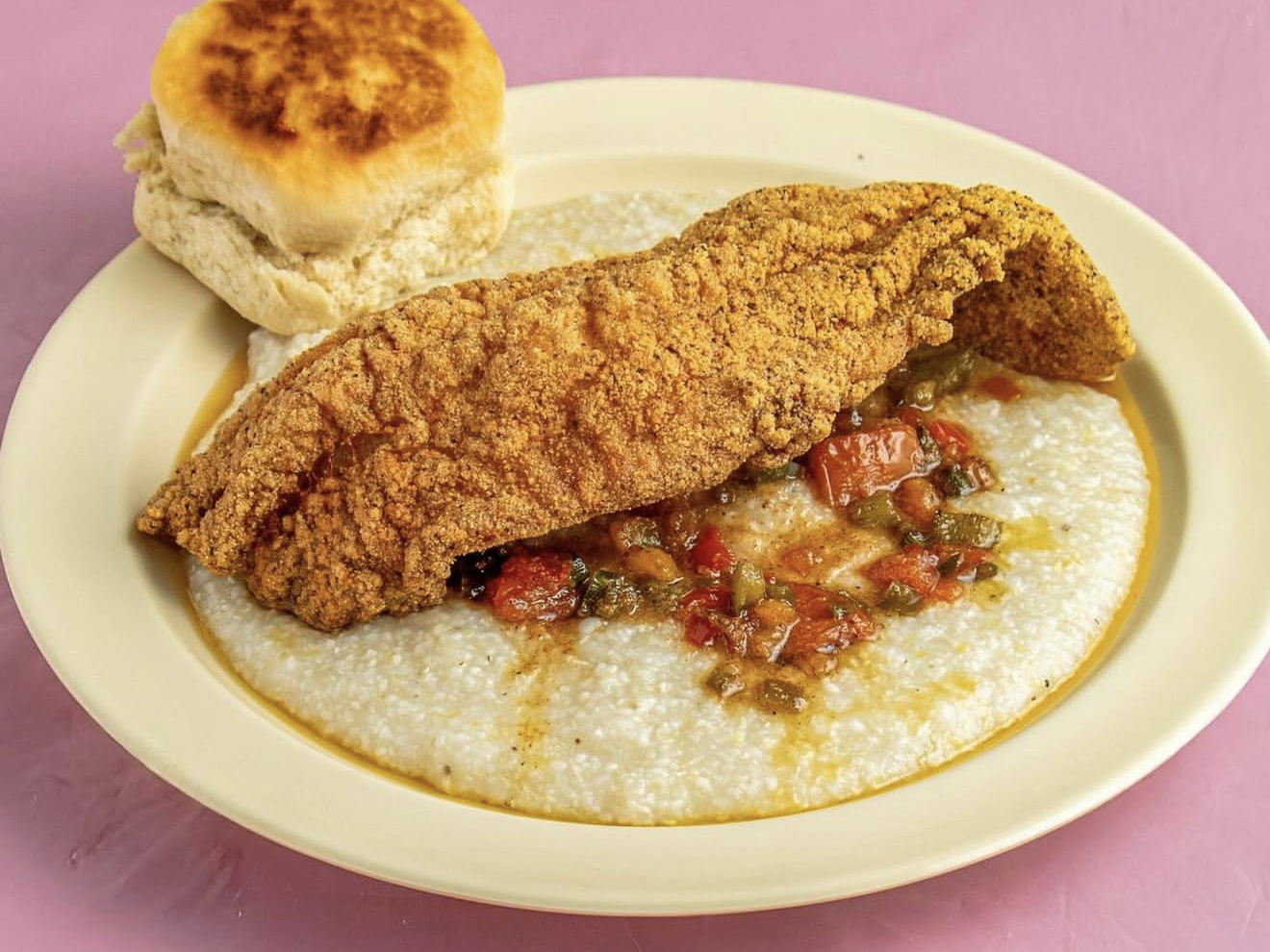a plate of grits with vegetables, fried catfish, and a biscuit on the side
