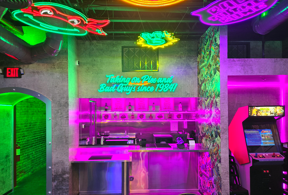 A Ninja Turtles themed bar with neon signs showing a Ninja Turtle and a sign that says "Taking in pies and bad guys since 1984!"