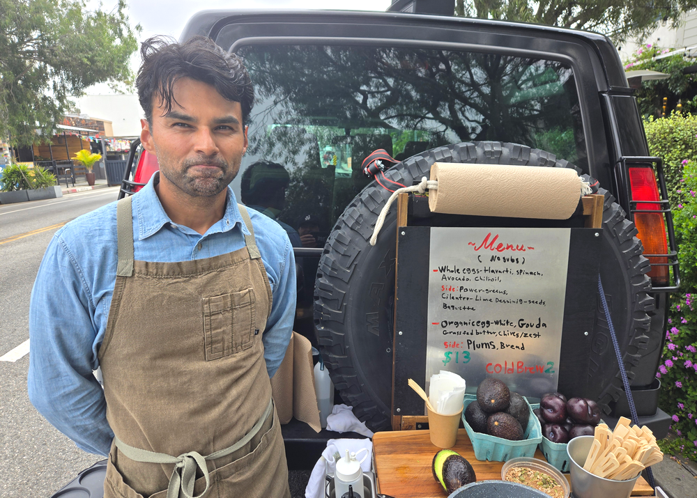 A man in an apron stands in front of his Land Rover, which doubles as a menu board for his street omelette business