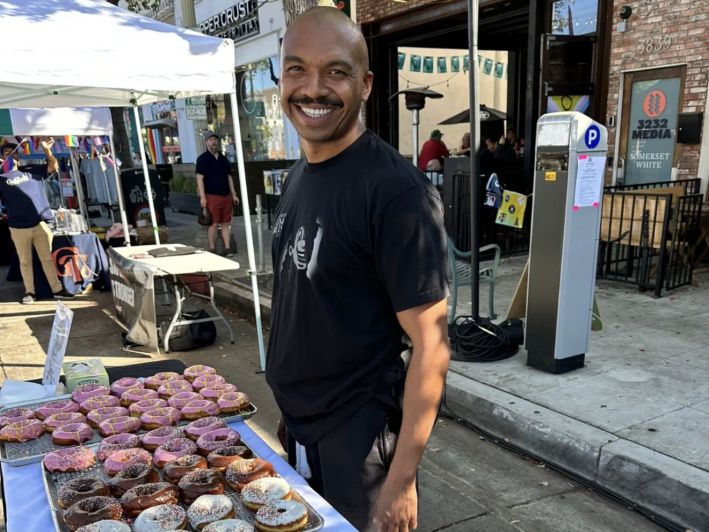a man smiles from behind a table filled with donuts