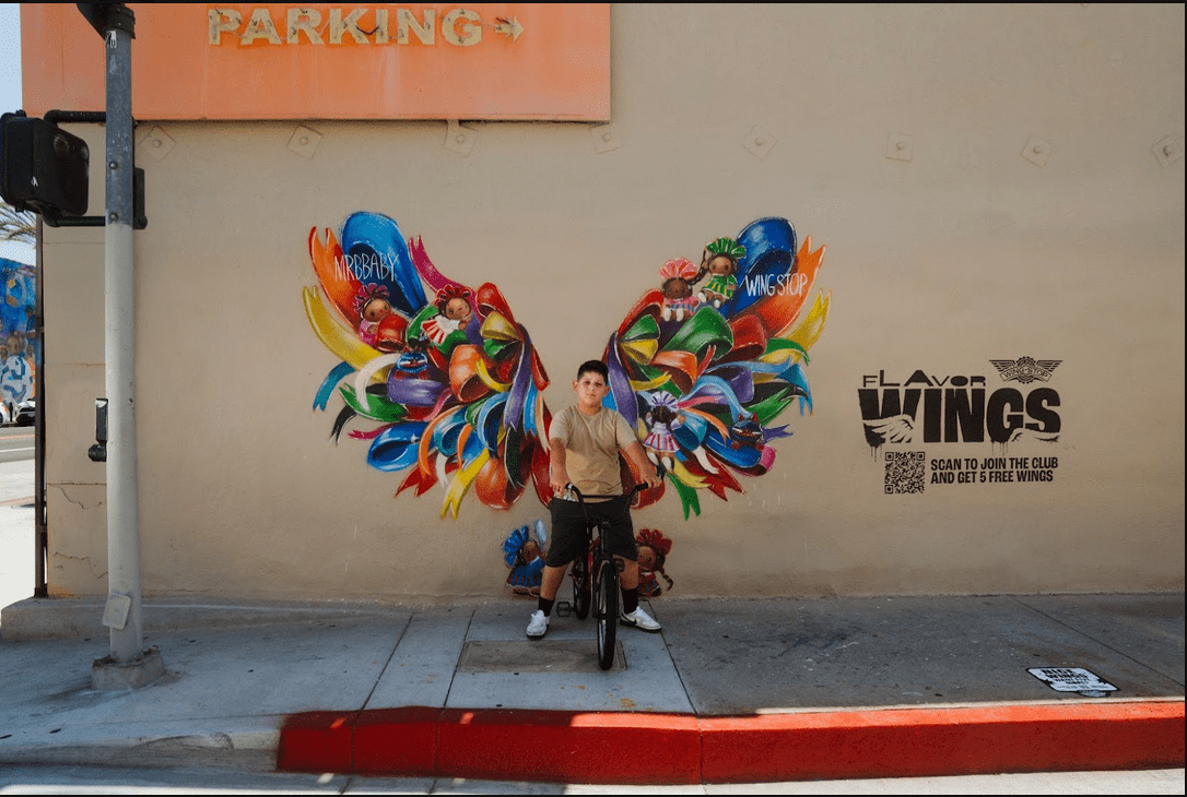 a youth on a bike poses before a giant set of painted wings festooned with bows, murals, and Maria dolls on an East L.A. street