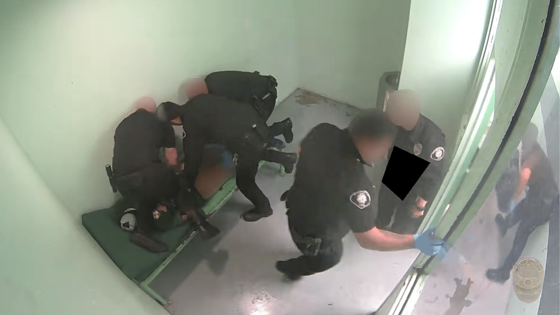a man lies on a jail bunk without pants with multiple officers around him, including one kneeling on him