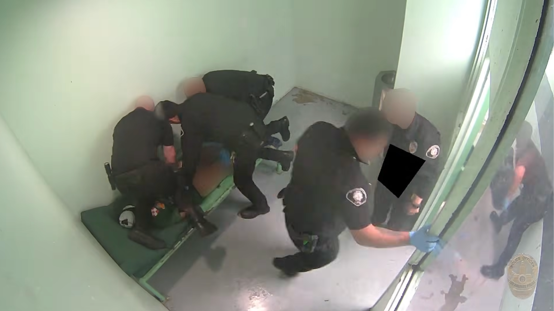 a man lies on a jail bunk without pants with multiple officers around him, including one kneeling on him
