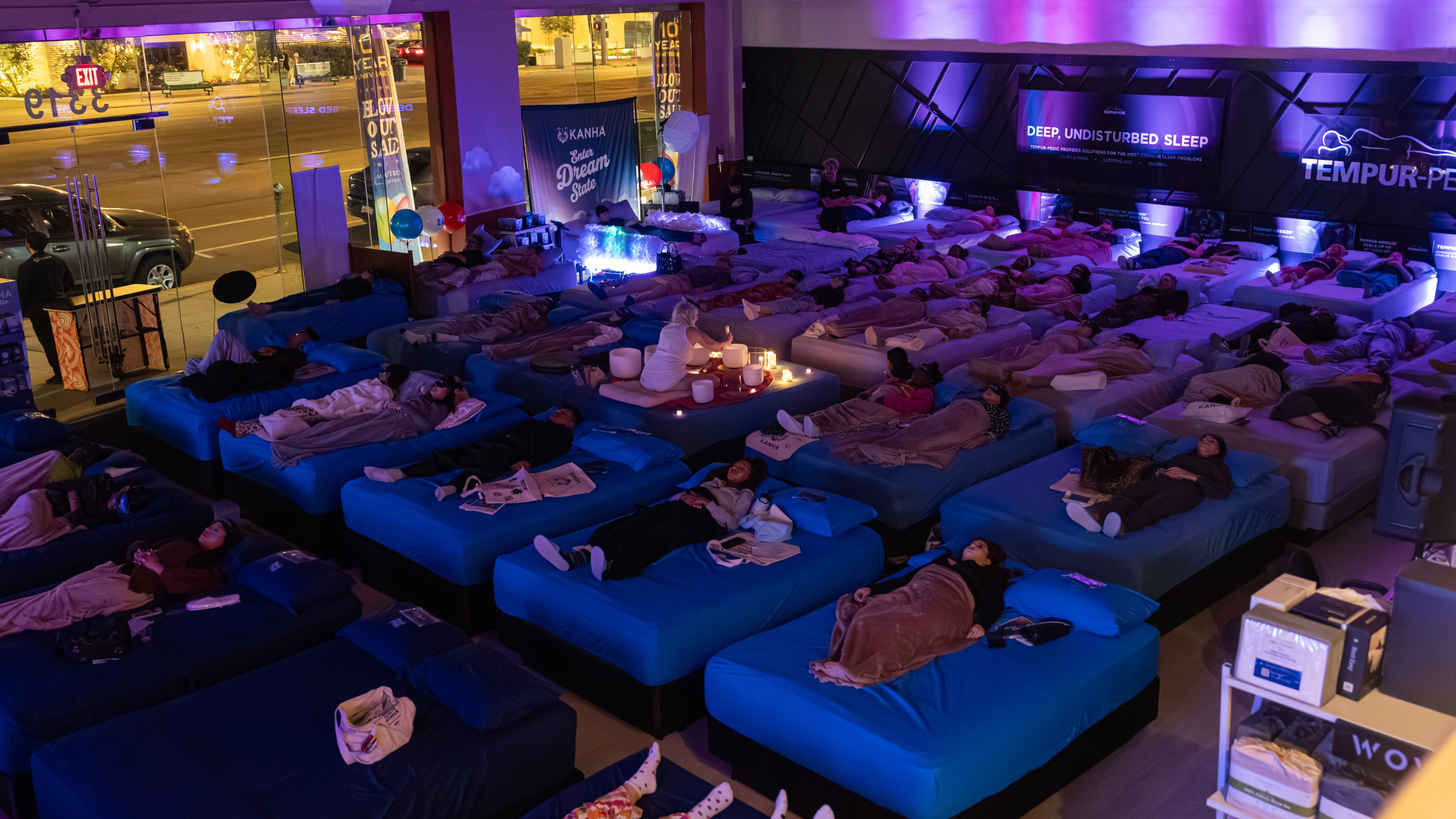 people relaxing on beds throughout a mattress store while a woman facilitates a sound bath