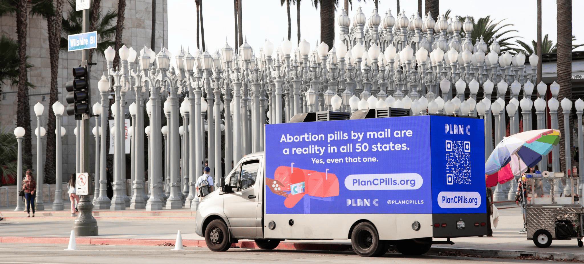 a truck with signage regarding abortion pills parks next to an outdoor art fixture, many tall lamp posts at LACMA