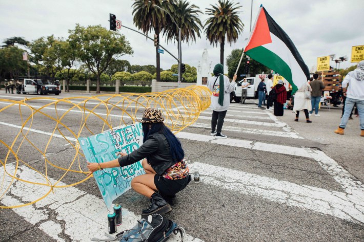 A protestor holding a sign kneels near a barricade next to two cans of spray paint.