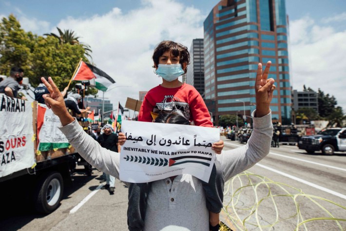 A young kid sitting on someone's shoulder with their hands raised in the air holding peace signs, holds a paper banner in front of their face.