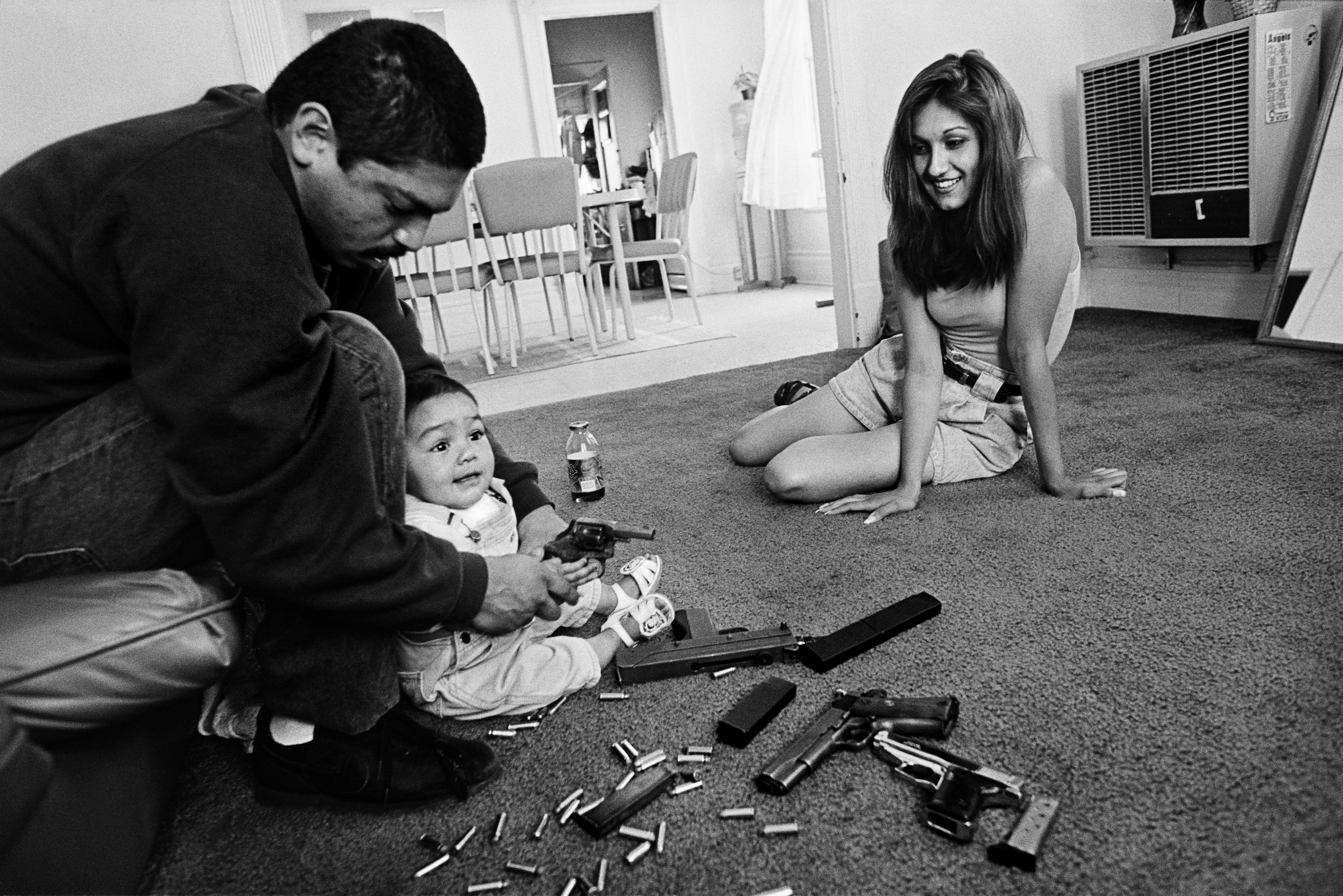 The morning after a rival gang tried to shoot Chivo for the fourth time. Chivo teaches his daughter how to hold a .32-caliber pistol. Her mother looks on. Boyle Heights, Los Angeles, CA, 1993